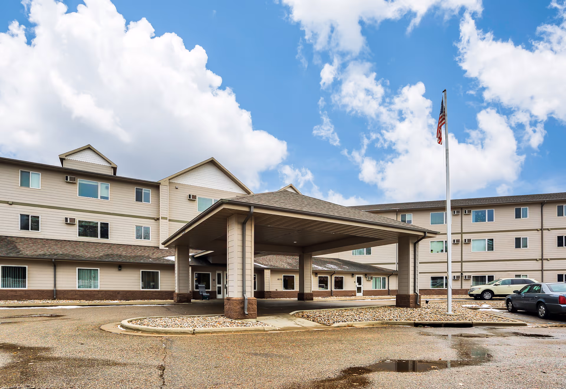 Exterior view of Minot Lodge Senior Living building with a covered entrance, multiple windows, parked cars, and an American flag on a flagpole under a partly cloudy sky.