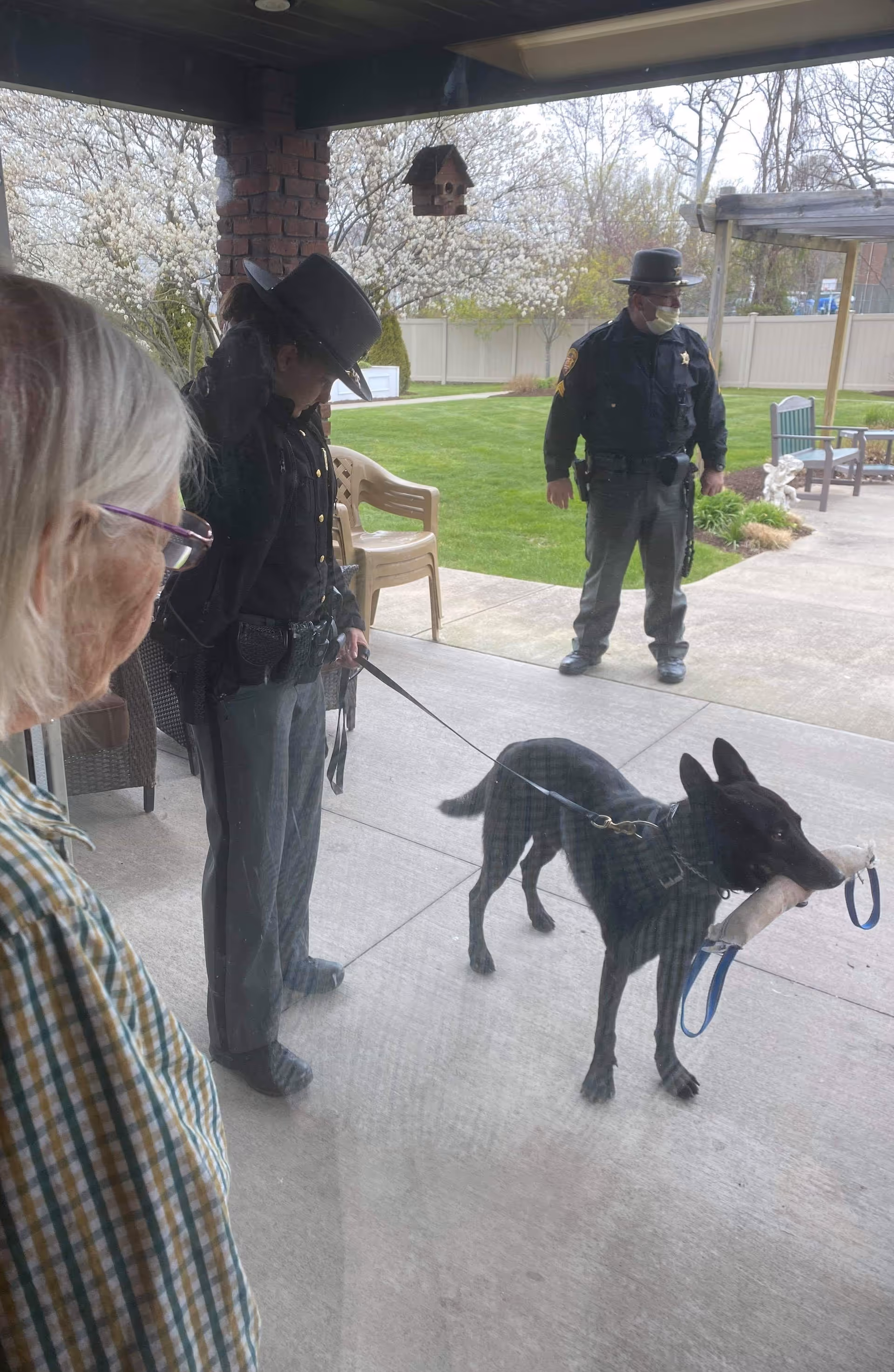 Two uniformed officers with hats stand outside on a patio area with a black dog on a leash holding a toy in its mouth. An elderly person with white hair and glasses looks on from inside through a glass door. The background shows a green lawn, trees with white blossoms, a birdhouse hanging from the patio roof, and outdoor chairs.