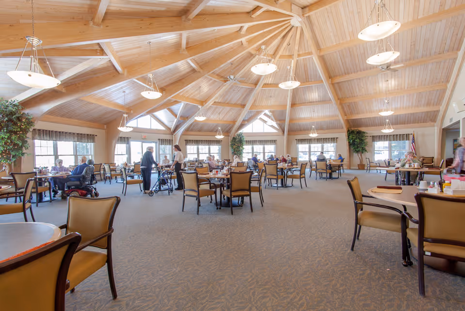 Spacious dining room in a retirement community with a high wooden vaulted ceiling, multiple round tables and chairs, large windows letting in natural light, and several elderly residents seated or walking with assistance.