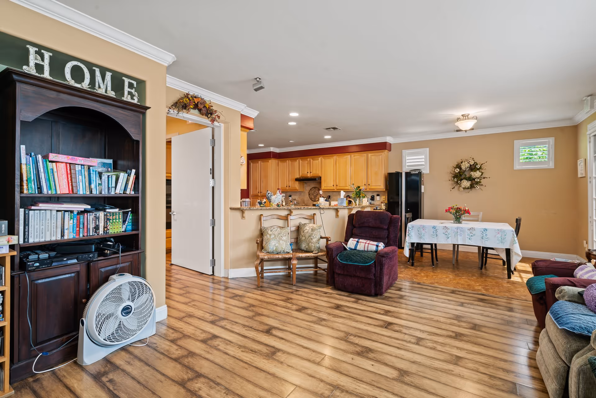 Interior view of a cozy senior living facility common area with wooden flooring, a dark wooden bookshelf filled with books and a fan in front, a pair of wooden chairs with cushions, a purple recliner chair, a dining table with floral tablecloth and flowers in a vase, and a kitchen area with light wooden cabinets and black refrigerator in the background.