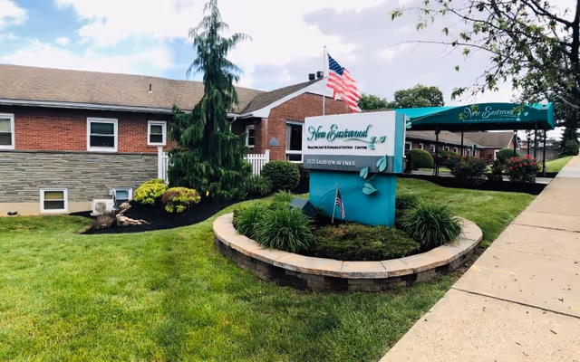 Exterior view of New Eastwood Healthcare And Rehabilitation Center showing a brick building with a green and white sign in front surrounded by landscaping. An American flag is flying above the sign, and there is a green awning over the entrance. The sky is partly cloudy.
