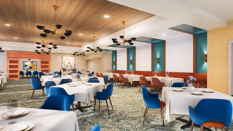 Bright, modern dining room with white-clothed tables, blue and red chairs, decorative chandeliers, and patterned carpet.