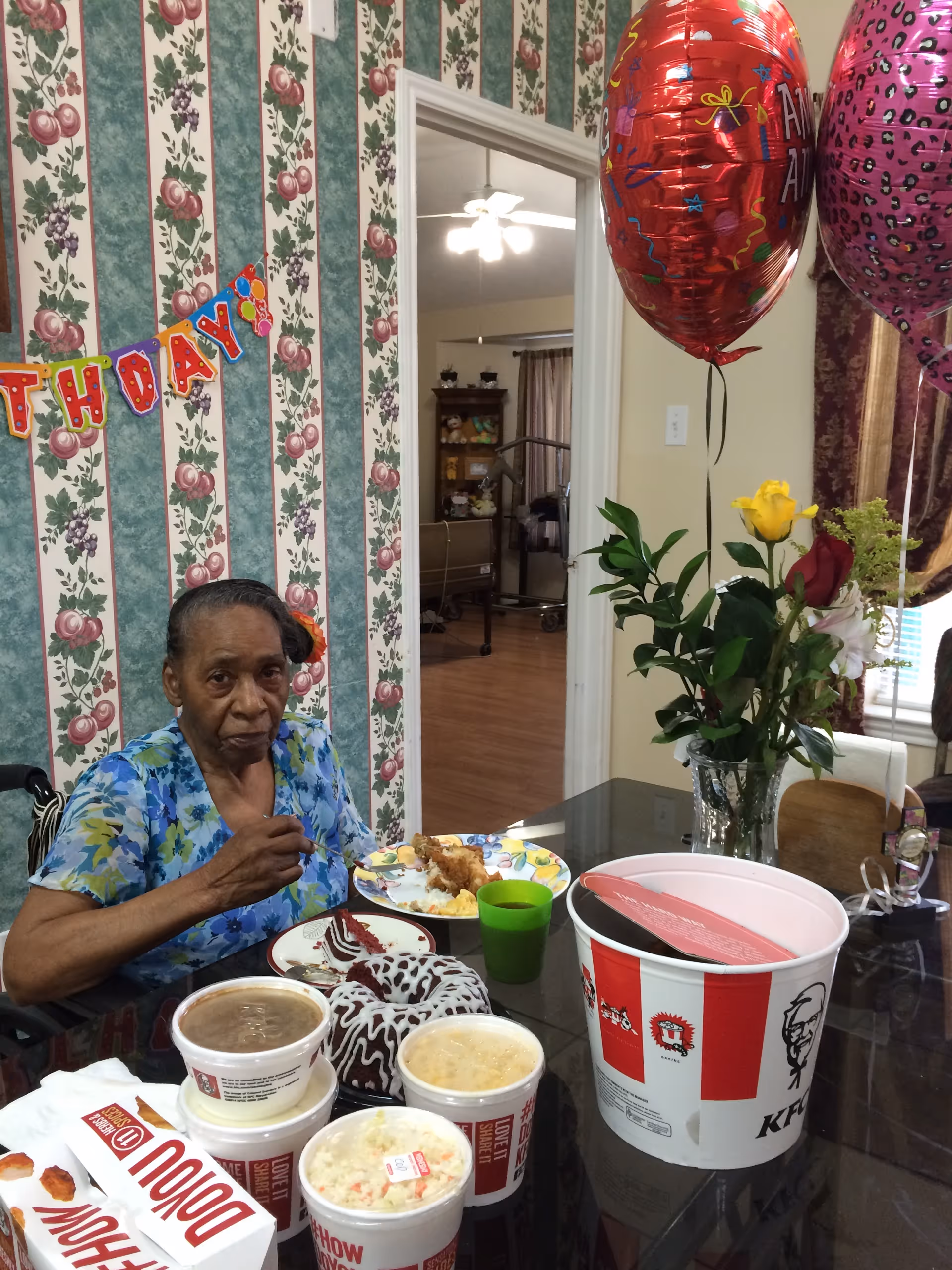 An elderly woman sits at a dining table with KFC food, cake, balloons and a vase of flowers in a decorated room.