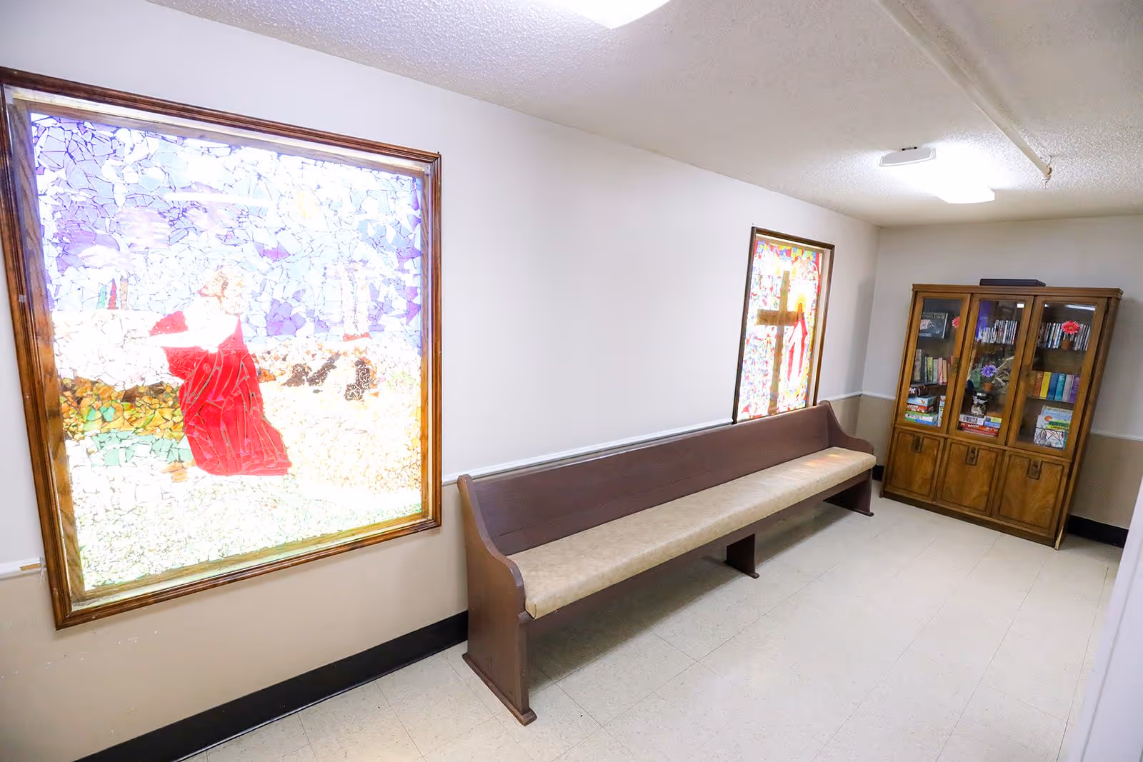 A hallway with two large stained glass windows on the left wall, a long wooden bench with a cushioned seat along the wall beneath the windows, and a wooden cabinet filled with books and decorative items at the end of the hallway.