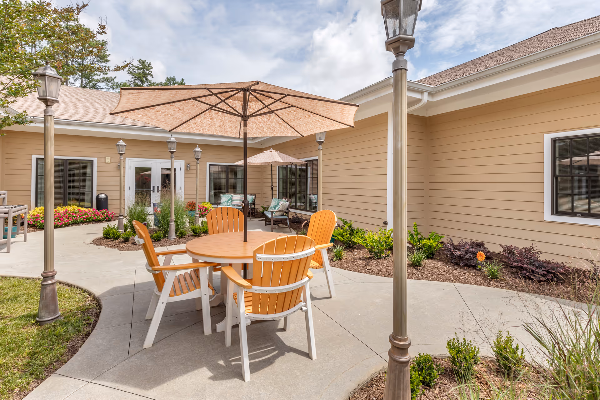 Outdoor courtyard patio with a round table, orange Adirondack chairs, umbrellas, lamp posts and landscaping in front of a beige building.
