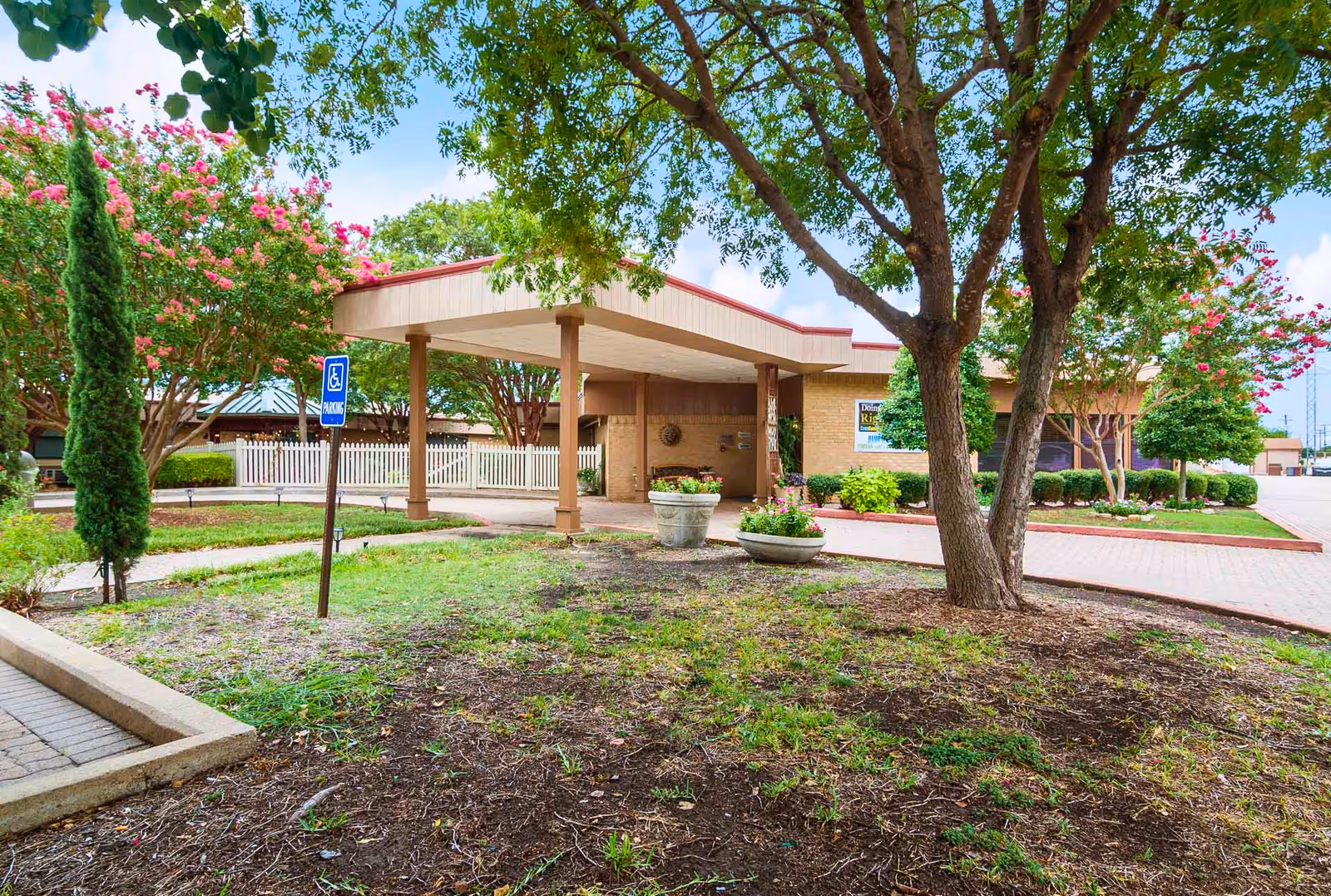 Covered entrance/porte-cochère of a senior living facility with trees, planters, and a handicapped parking sign.