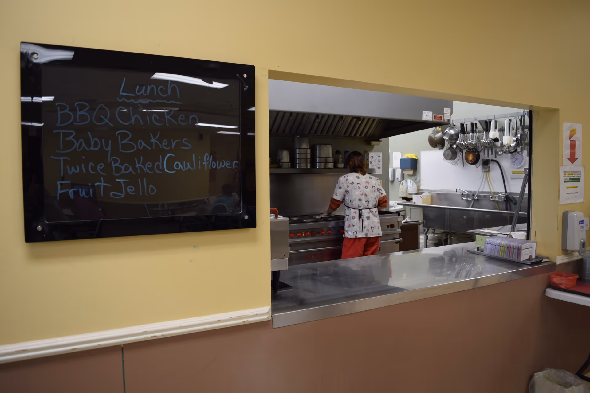 View through a serving window into a kitchen where a person in a white and red uniform is cooking. To the left of the window, a blackboard displays the lunch menu: BBQ Chicken, Baby Bakers, Twice Baked Cauliflower, and Fruit Jello. Various kitchen utensils hang on the wall inside the kitchen.