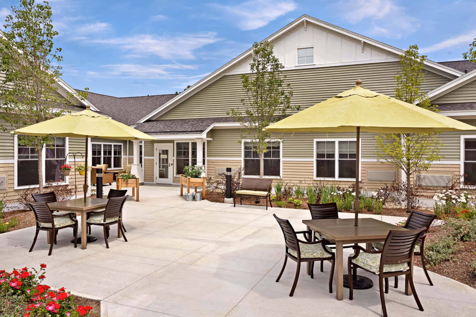 Outdoor patio area at Avita of Wells featuring two square tables each with four chairs and large yellow umbrellas. The patio is surrounded by small trees, flower beds, and a beige and green building with multiple windows and a door.
