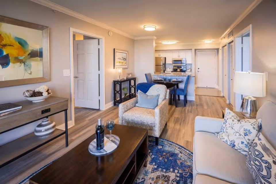A bright and modern living room and dining area in an apartment. The living room features a beige sofa with patterned cushions, a patterned armchair with a blue throw, a wooden coffee table with a bottle of wine and two glasses, and a decorative console table with artwork above it. The dining area has a round table set for four with blue upholstered chairs. The kitchen in the background includes stainless steel appliances and white cabinetry. The space has wood flooring and neutral-colored walls with ceiling lights.