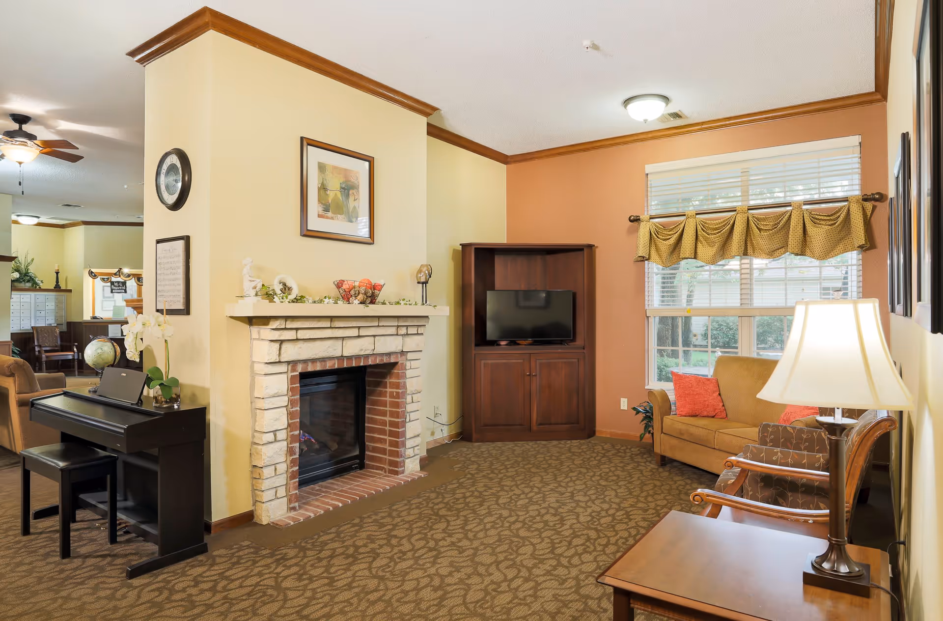 A cozy living room area in an assisted living facility featuring a stone fireplace with decorative items on the mantel, a wooden TV cabinet with a flat-screen TV, a brown sofa with red cushions, an armchair, a side table with a lamp, a piano with a bench, and a large window with a valance allowing natural light to enter.