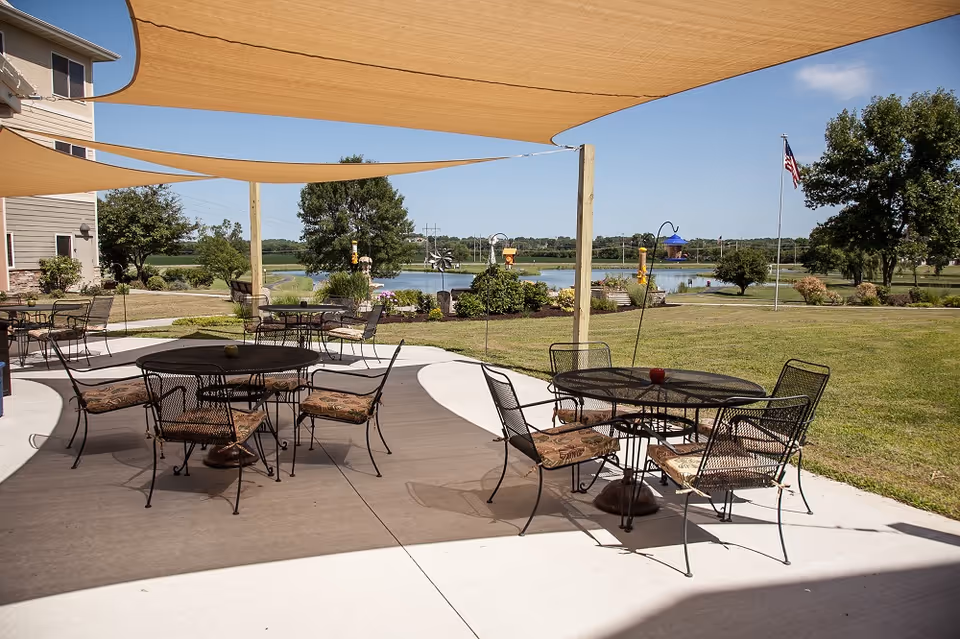 Outdoor patio area with several round metal tables and cushioned chairs under beige shade sails. The patio overlooks a grassy lawn with trees, a small pond, and an American flag on a flagpole. The sky is clear and blue.