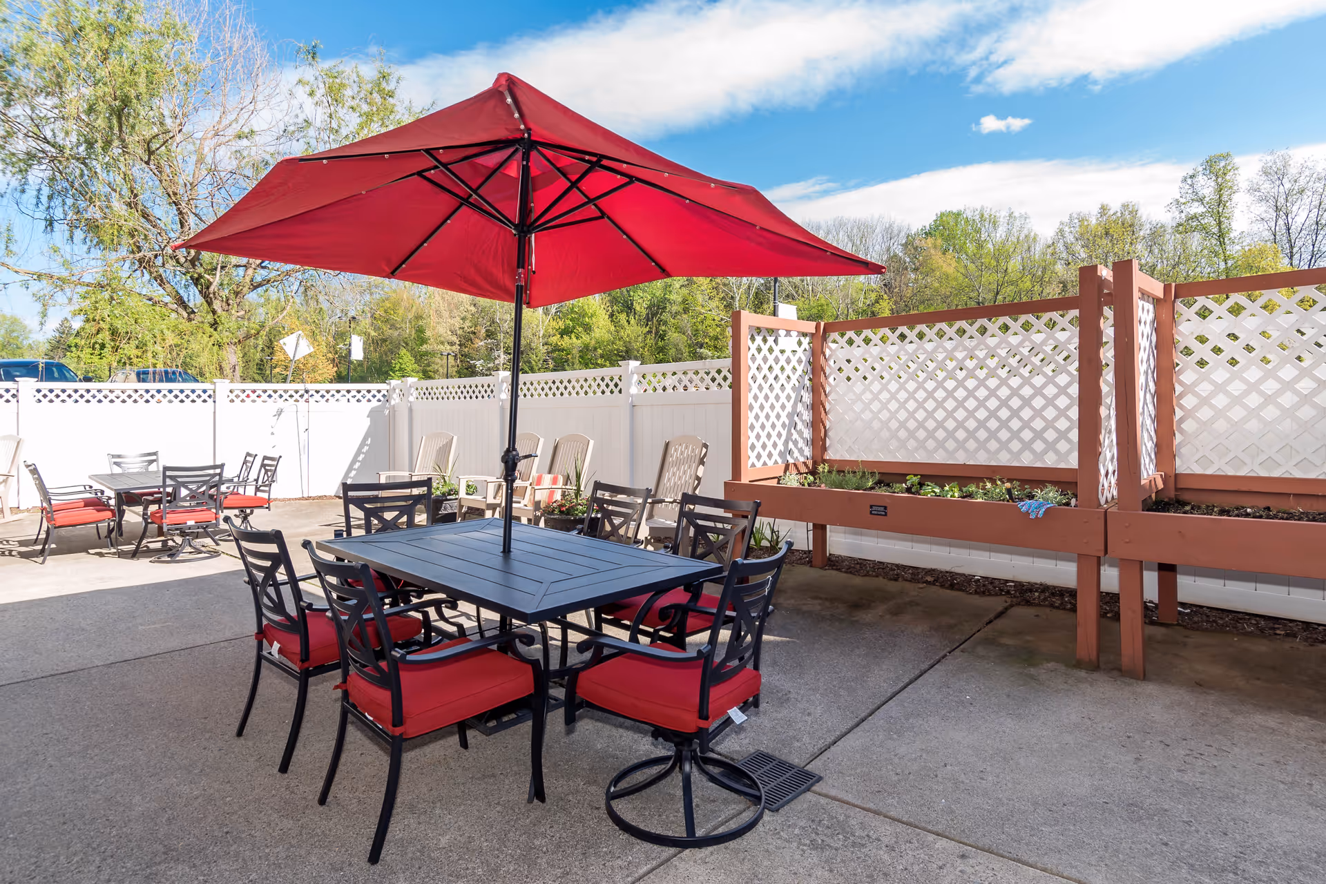 Outdoor patio area with black metal tables and chairs featuring red cushions. One table has a large red umbrella providing shade. Raised garden beds with lattice fencing are visible along the side, and trees and a blue sky with some clouds are in the background.