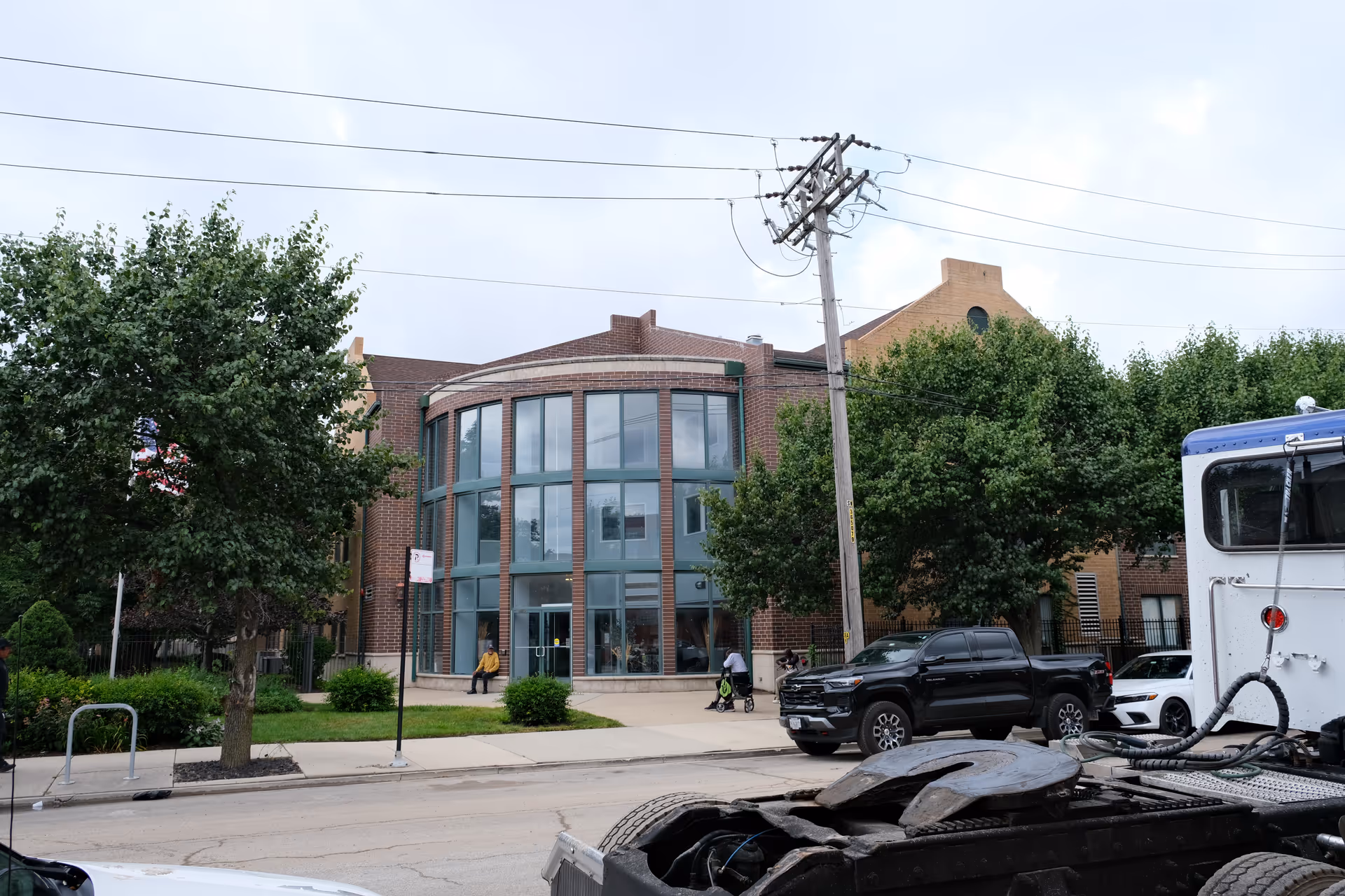 Exterior view of a brick building with large curved glass windows and a sidewalk in front. There are trees and parked vehicles along the street, including a black pickup truck and a white vehicle. A person is sitting on a bench near the entrance, and another person is pushing a stroller on the sidewalk.