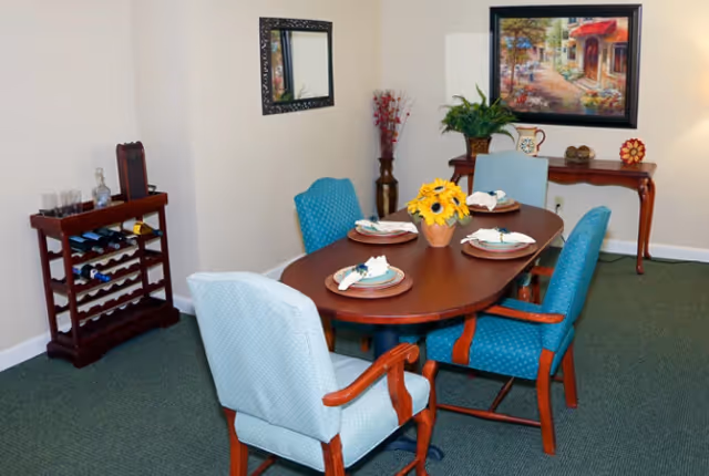 A dining room with a wooden oval table set for four with plates, napkins, and a centerpiece of yellow flowers. Four upholstered chairs surround the table. A wooden wine rack with bottles and glasses is against the wall on the left. A mirror hangs on the wall above the wine rack. On the right wall, there is a framed painting above a wooden console table decorated with a plant and other small items.