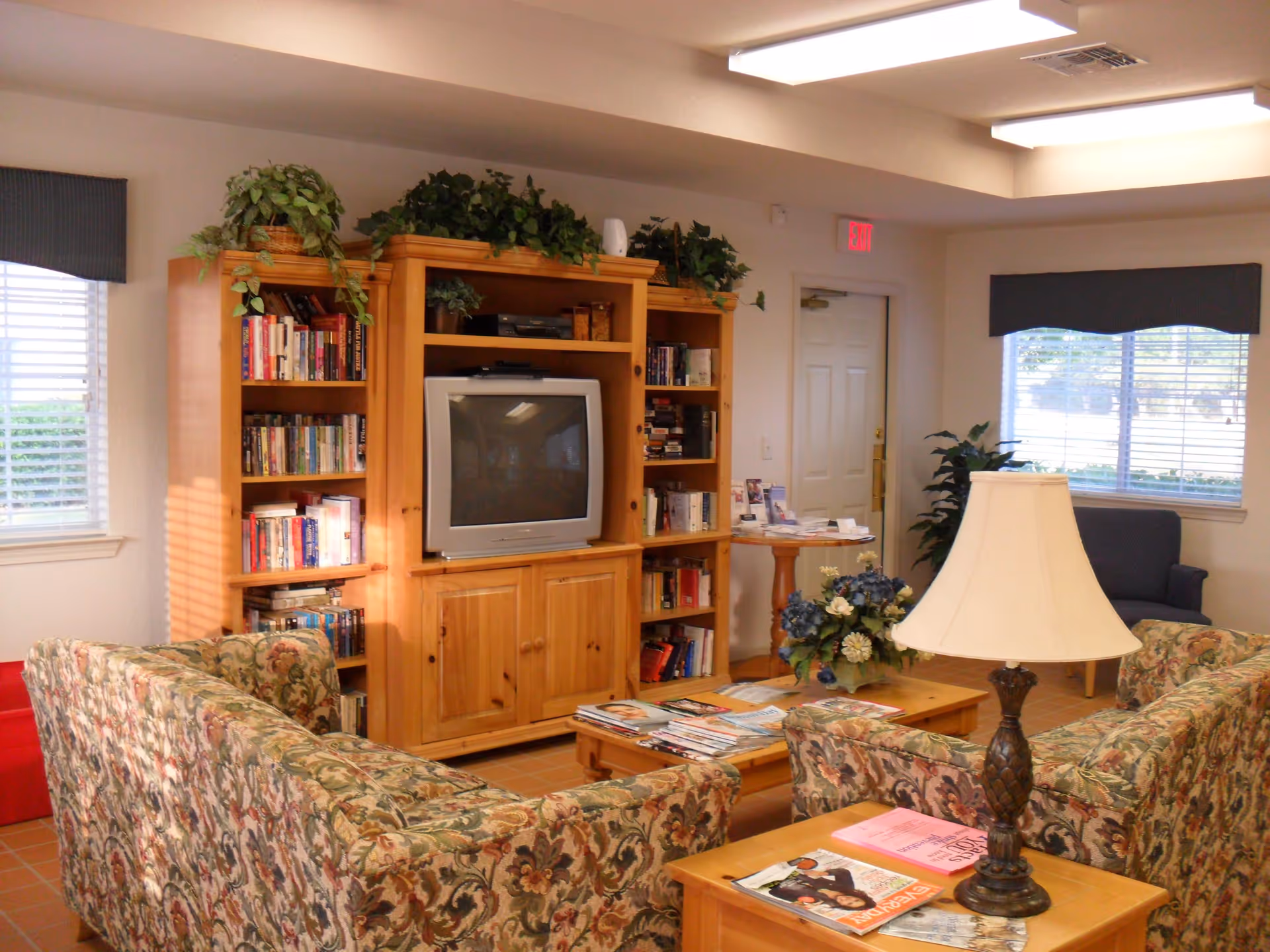 A cozy living room with floral patterned sofas arranged around a wooden coffee table holding magazines and a flower arrangement. A wooden entertainment center with bookshelves and a TV is against the wall. There are windows with blinds and valances, a table with brochures near a door, and a lamp on a side table.