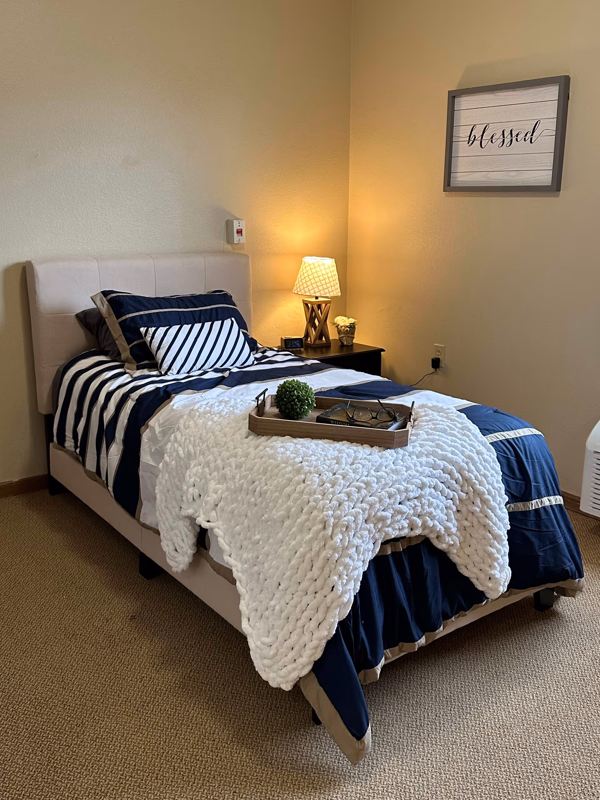 A neatly made single bed with navy blue and white striped bedding and a thick white knitted blanket draped over the foot of the bed. On the bed is a wooden tray holding a small green plant, a book, and a pair of glasses. Next to the bed is a dark wooden nightstand with a lamp, a small flower pot, and a digital clock. The walls are beige, and a framed sign with the word 'blessed' hangs above the nightstand.