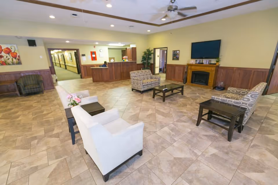 A spacious and well-lit common area in a senior living facility featuring several armchairs and sofas arranged around coffee tables on a tiled floor. There is a wooden reception desk in the background with a person seated behind it, a hallway leading further into the building, a wall-mounted flat screen TV above a wooden fireplace, and some decorative plants and artwork on the walls.