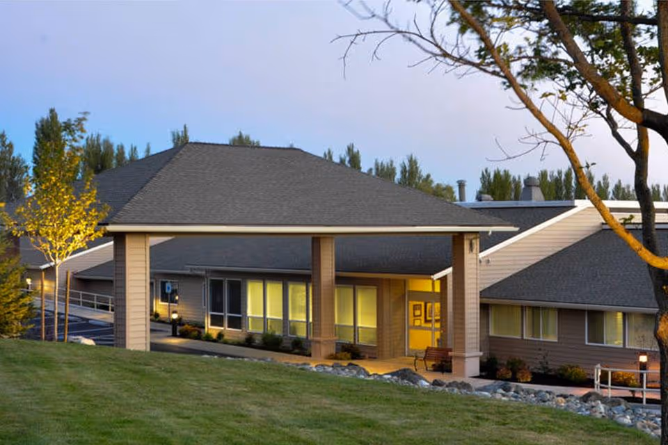 Exterior view of a single-story building with a covered entrance supported by columns, surrounded by grass and trees during early evening with lights on inside the building.