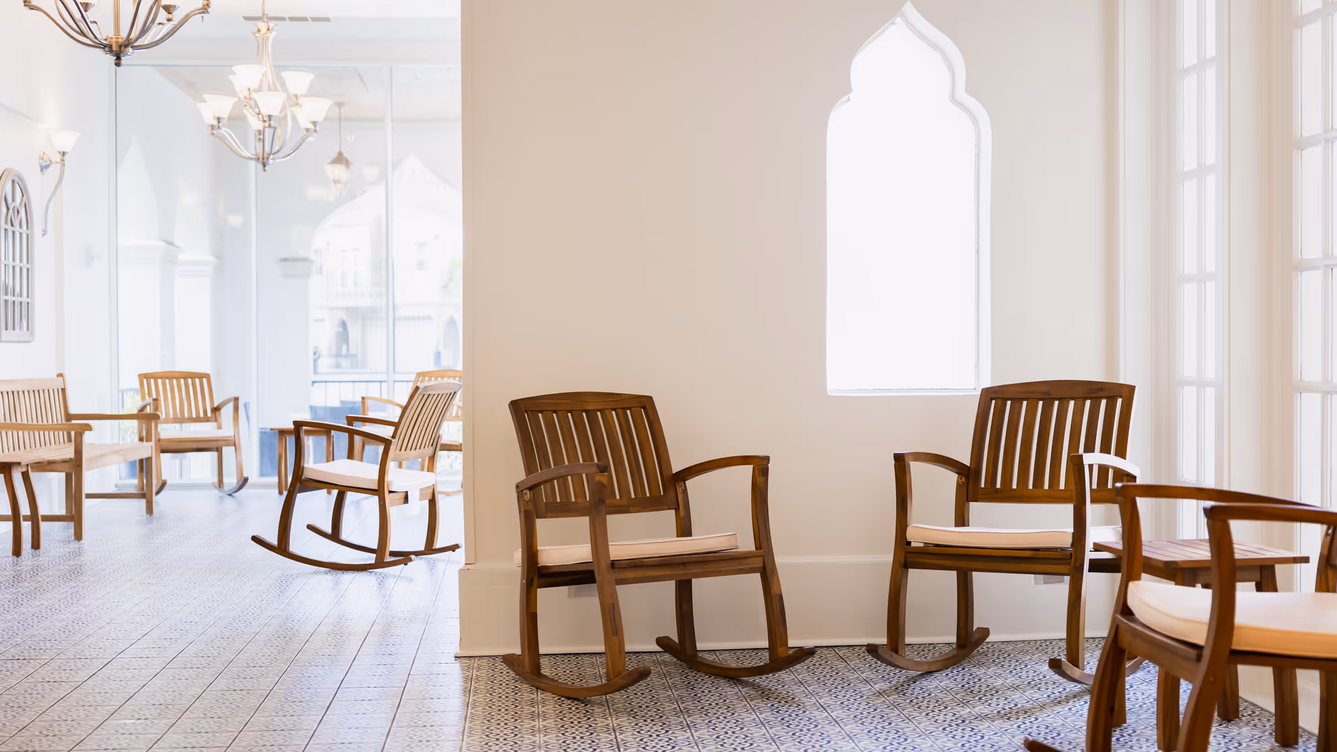 A bright and airy common area with wooden rocking chairs and armchairs arranged on a patterned tile floor. The room features large windows and decorative light fixtures, creating a welcoming and comfortable atmosphere.