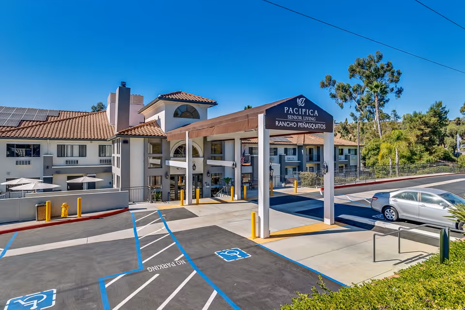 Exterior view of Pacifica Senior Living Rancho Penasquitos building with a covered entrance, parking spaces including handicapped spots, and surrounding greenery under a clear blue sky.