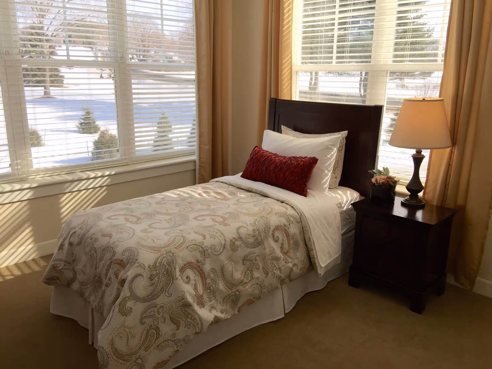 A cozy bedroom with a single bed featuring a paisley-patterned comforter, white sheets, and a red decorative pillow. Next to the bed is a dark wooden nightstand with a lamp and a small plant. Large windows with white blinds and beige curtains let in natural light, showing a snowy outdoor scene.