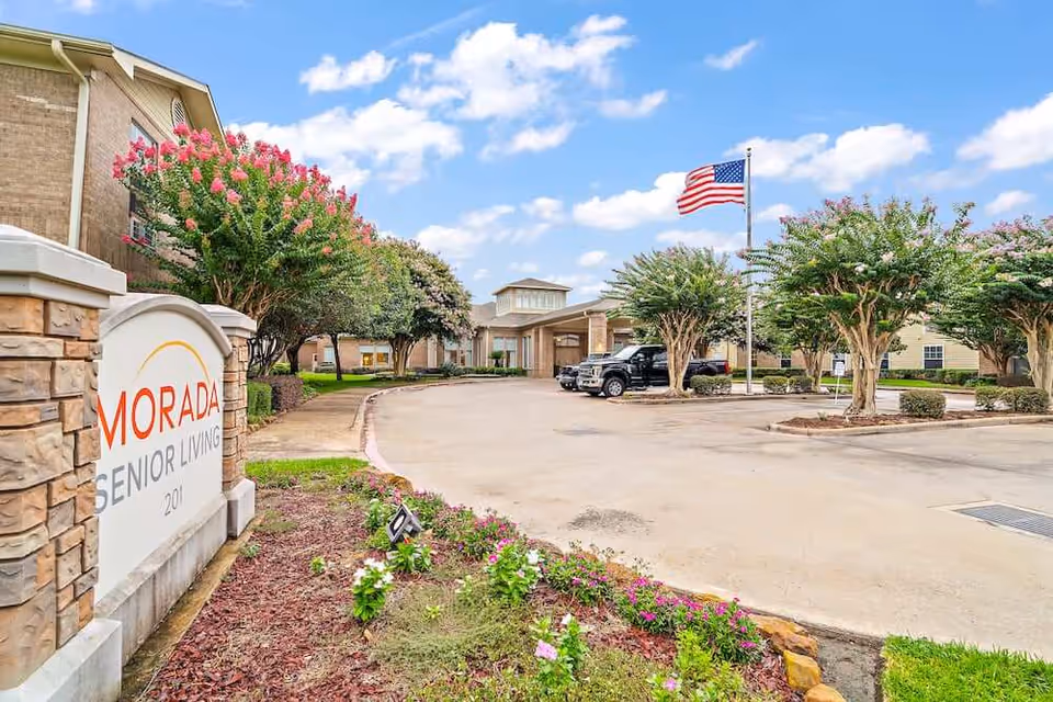 Exterior view of Morada Senior Living facility with a stone sign in the foreground, a driveway leading to the entrance, several trees with pink flowers, an American flag on a flagpole, and a partly cloudy sky.