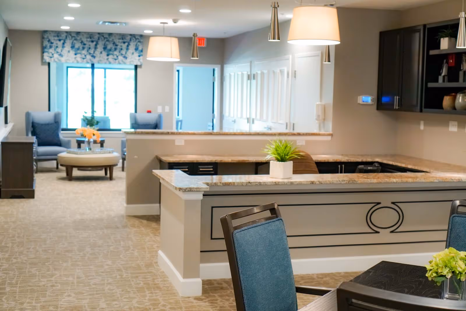 Interior view of a senior living facility common area featuring a granite countertop reception desk with a small potted plant, blue upholstered chairs around a dark wooden table, two blue armchairs near a window with floral valance, and soft overhead lighting.