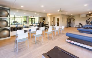 Bright exercise room with treadmills and other fitness equipment, a row of white chairs facing a mirrored wall, and large windows letting in natural light.