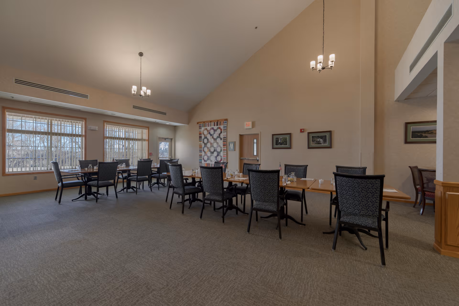 A spacious dining room with multiple tables and chairs arranged neatly. The room has large windows with vertical blinds allowing natural light to enter. The walls are decorated with framed pictures and a quilt hanging near an exit door. The ceiling is high with two modern chandeliers providing light.