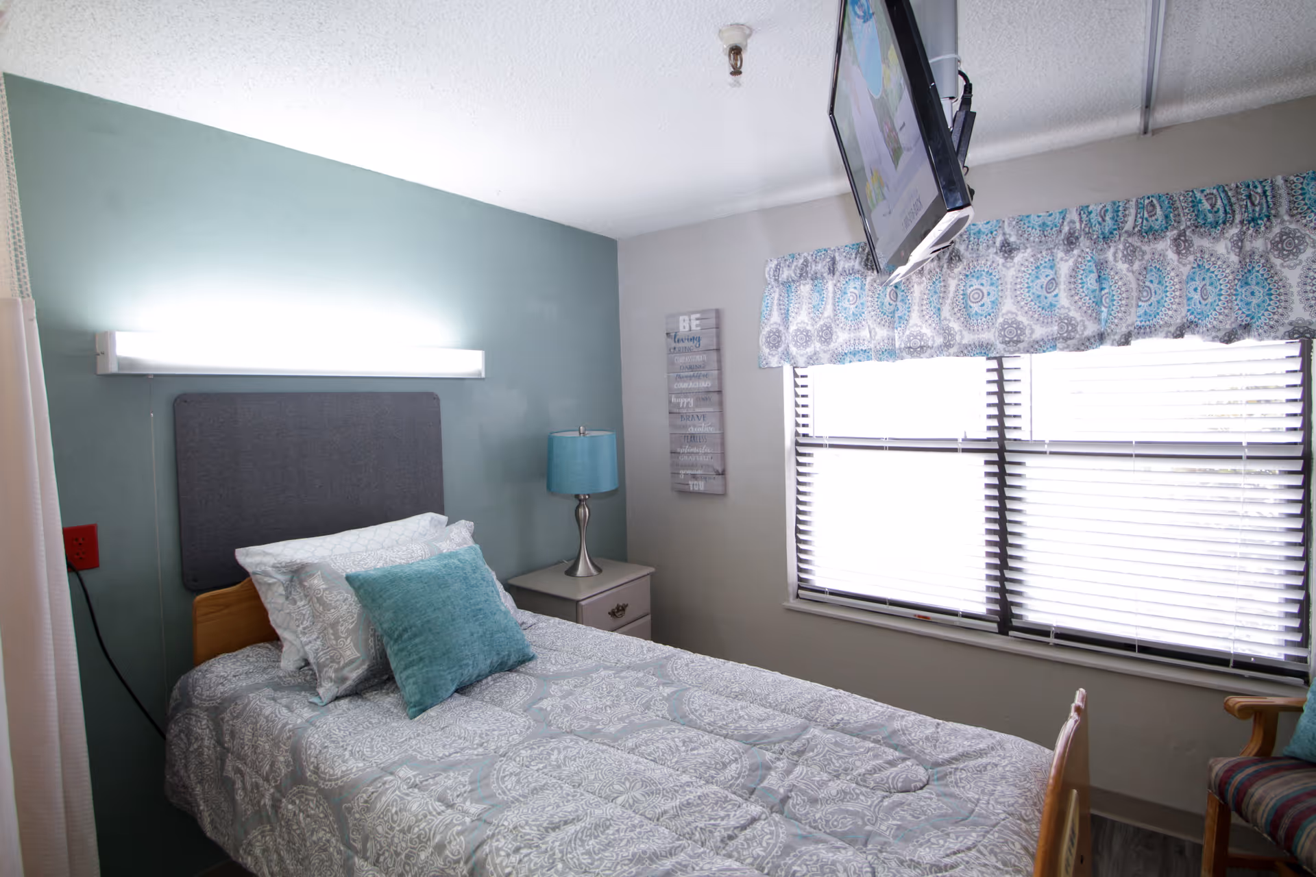 A small bedroom with a single bed featuring patterned bedding and pillows. There is a nightstand with a blue lamp next to the bed. A window with blinds and a patterned valance lets in natural light. A wall-mounted TV is positioned above the window, and a decorative sign hangs on the wall.