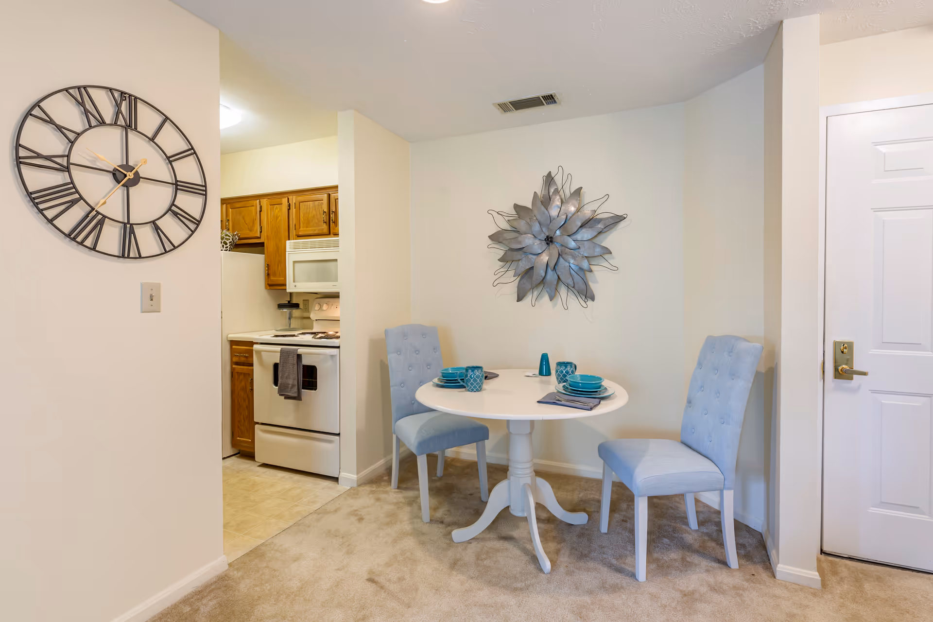 Small dining nook with a round white table and two light-blue chairs, a decorative wall sculpture, and a view into a compact kitchen.