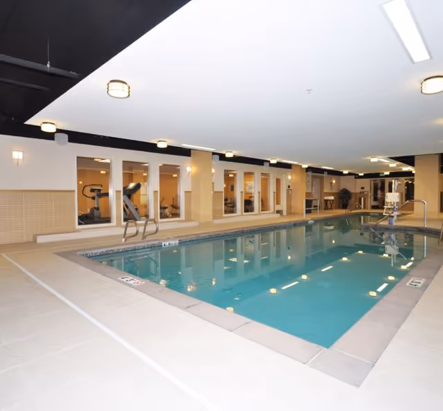 Indoor swimming pool with clear water surrounded by beige tiled flooring. The pool area is well-lit with ceiling lights and has a pool lift for accessibility. Behind the pool, large windows reveal a fitness room with exercise equipment such as treadmills and stationary bikes.
