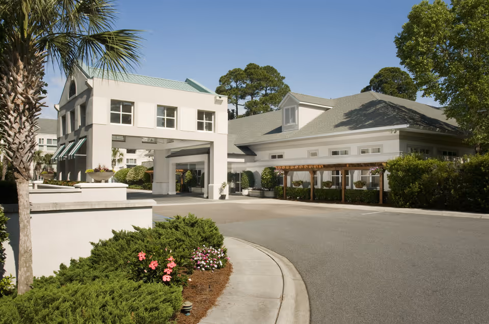 Exterior view of a senior living facility with a driveway, landscaped bushes, flowers, palm trees, and a large building with a covered entrance and multiple windows under a clear blue sky.