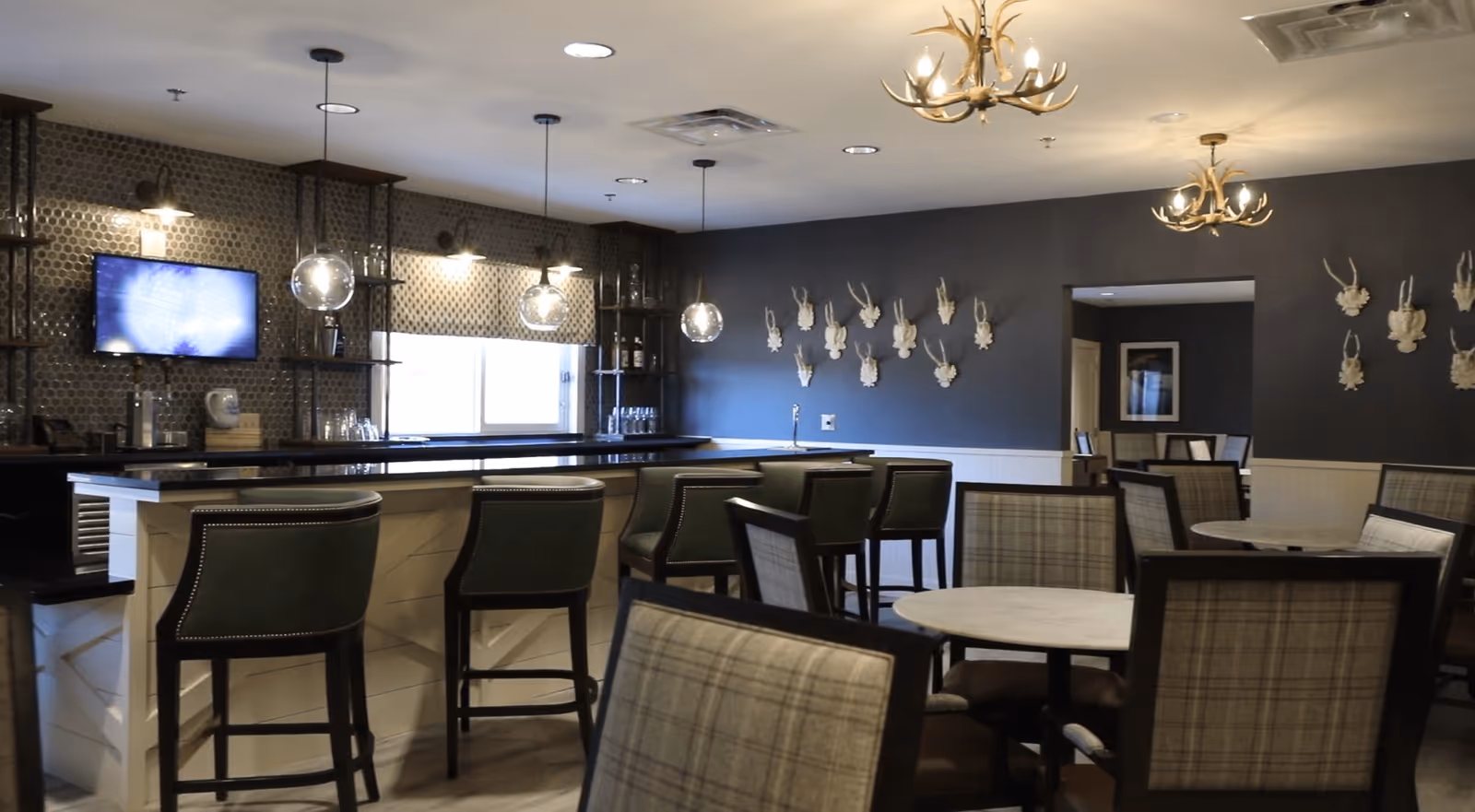 Communal dining and bar area with high stools, round tables and plaid chairs, pendant lights and decorative wall-mounted antler sculptures.