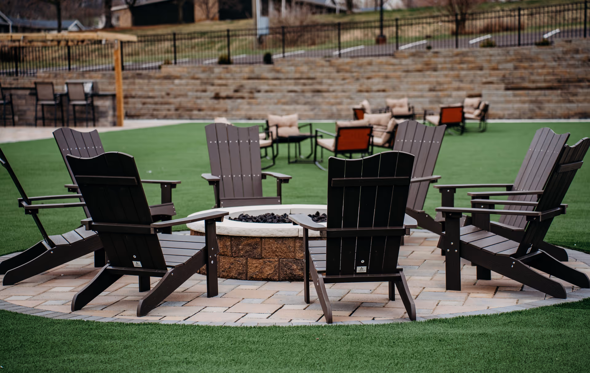 Outdoor seating area with dark wooden Adirondack chairs arranged around a circular fire pit on a paved surface, surrounded by artificial green turf. In the background, there are additional cushioned chairs and tables, a stone retaining wall, and a metal fence.