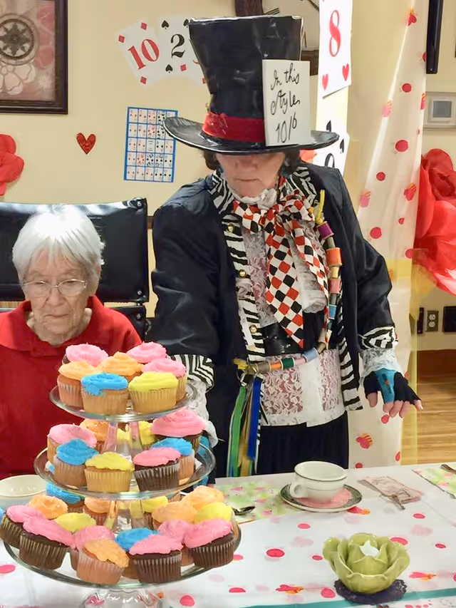 An elderly woman sitting at a table with a tiered tray of colorful frosted cupcakes. Next to her stands a person dressed as the Mad Hatter from Alice in Wonderland, wearing a large black top hat with a card that reads 'In this style 10/6', a patterned scarf, and a colorful outfit. The table is decorated with a floral tablecloth and a teacup, and the background has playing card decorations on the wall.