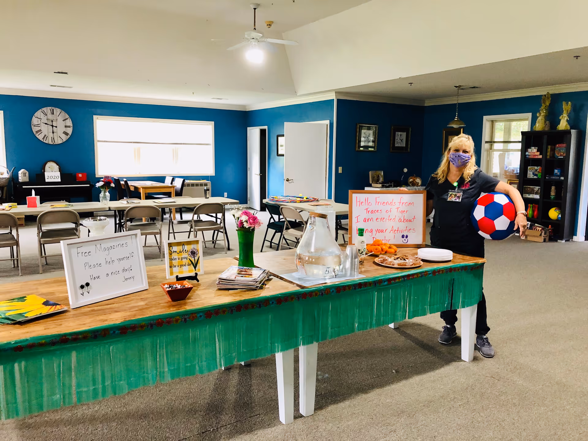 A spacious activity room with blue walls and several tables and chairs arranged for group activities. A woman wearing a face mask stands near a long table covered with a green cloth, holding a large red, white, and blue ball. The table has a pitcher of water, plates with snacks, flowers, and signs including one that reads 'Free Magazines Please help yourself Have a nice day! Jenny' and another welcoming friends from Traces of Tiger Retirement and expressing excitement about upcoming activities.