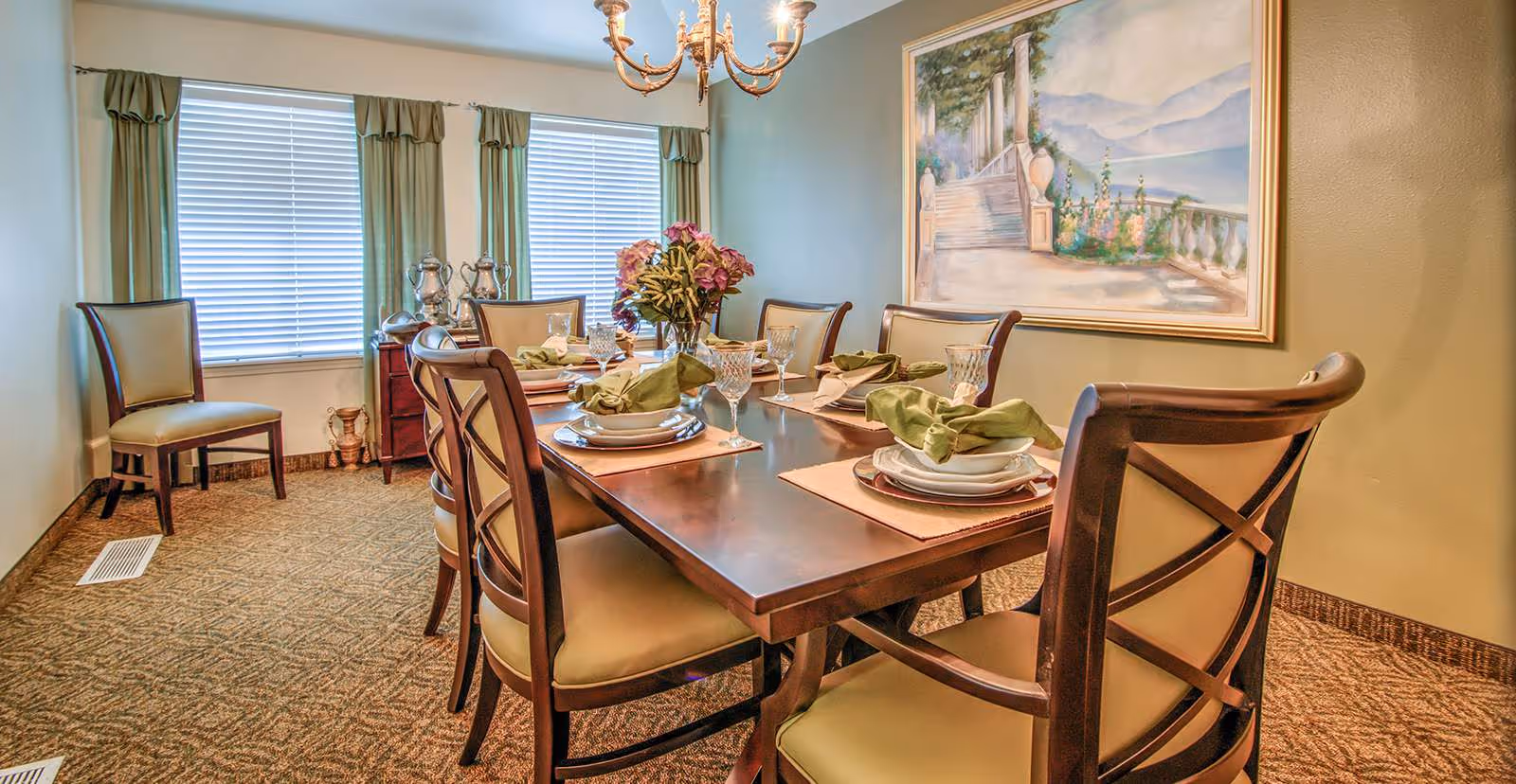A dining room with a wooden table set for six people, featuring plates, glasses, and green napkins. The room has carpeted flooring, two windows with green curtains, a chandelier overhead, and a large framed painting on the wall depicting a scenic outdoor staircase with mountains in the background.
