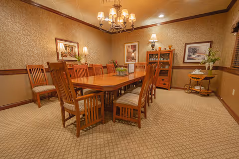 A warmly lit dining room with a long wooden table surrounded by wooden chairs with cushions. The room has patterned wallpaper, framed artwork on the walls, a chandelier hanging from the ceiling, and a wooden cabinet with decorative items. There is also a small side table with flowers and a lamp.