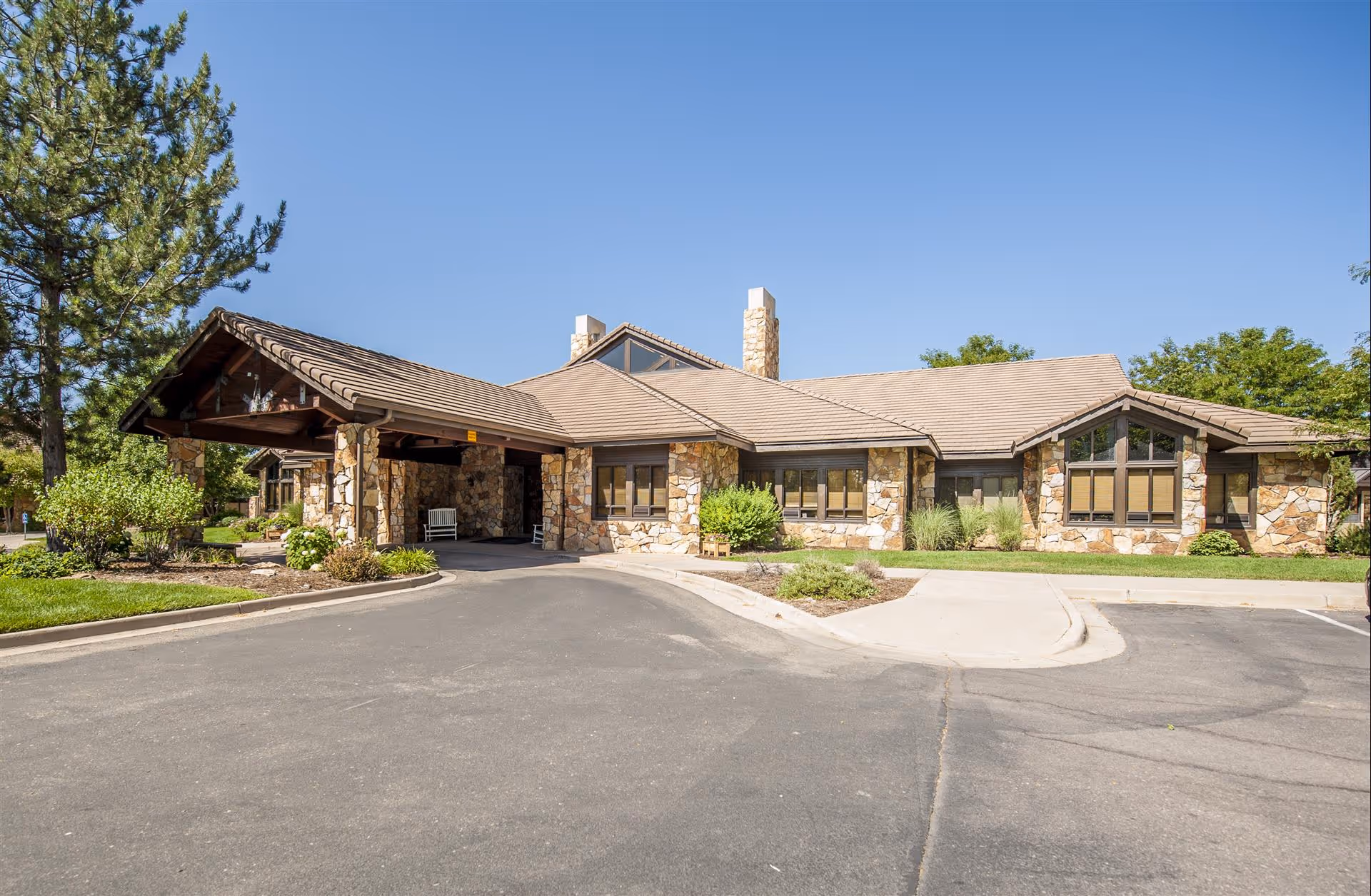 Exterior view of a single-story assisted living facility building with stone walls, a covered entrance, and a paved driveway. The building is surrounded by greenery including trees, bushes, and a well-maintained lawn under a clear blue sky.