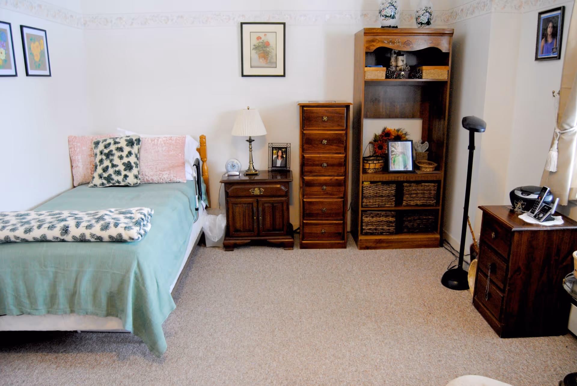 A cozy bedroom with a single bed covered in a green blanket and floral pillows. Next to the bed is a wooden nightstand with a lamp, a clock, and a framed photo. There is a tall wooden dresser and a wooden shelving unit with baskets and decorative items. The room has light-colored walls with framed pictures and carpeted flooring.