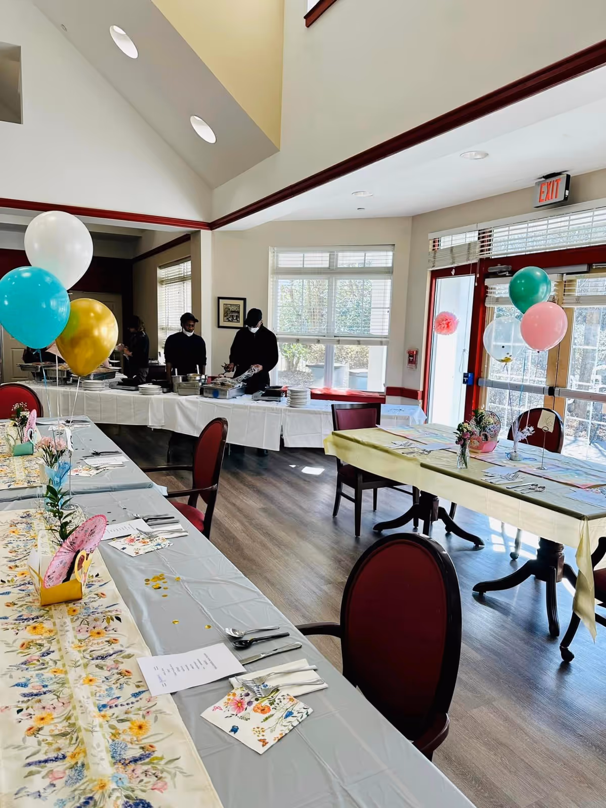 A decorated dining room with tables set for a meal, featuring floral table runners, colorful balloons, and neatly arranged utensils and napkins. Three people are serving food from a buffet table in the background near large windows letting in natural light.