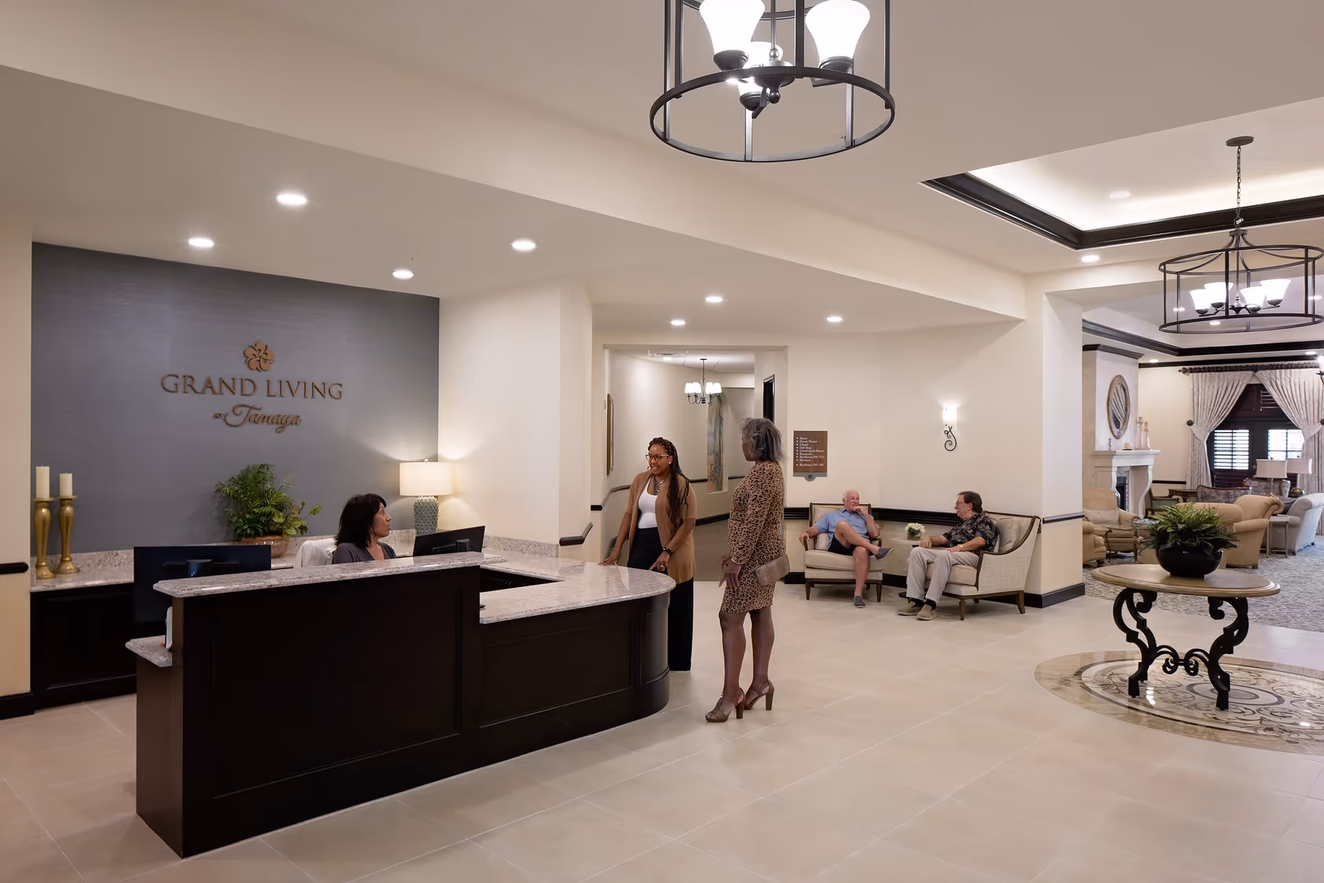 Reception area of Grand Living at Tamaya with a front desk where a woman is seated, two women standing and talking, and two men sitting on chairs in the background. The space is well-lit with modern light fixtures and has a welcoming, elegant interior design.
