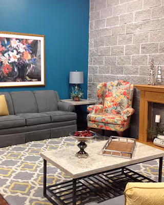 A cozy living room with a gray sofa, a floral patterned armchair, a wooden fireplace, and a coffee table with decorative items. The walls are painted teal and gray brick, and there is a colorful floral painting hanging above the sofa.