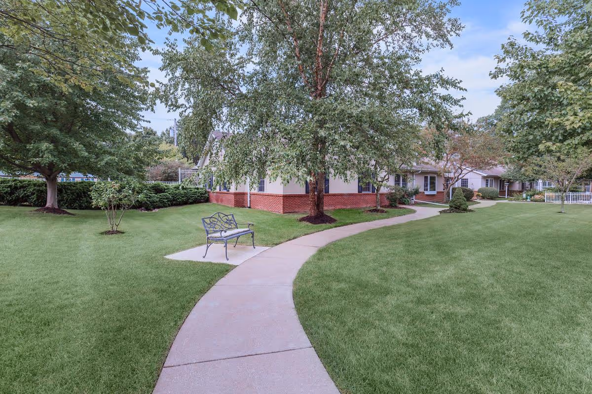 A winding concrete pathway through a well-maintained grassy area with trees and shrubs, leading to a single-story building with a brick foundation and light-colored walls. There is a decorative metal bench on a small concrete pad beside the path.