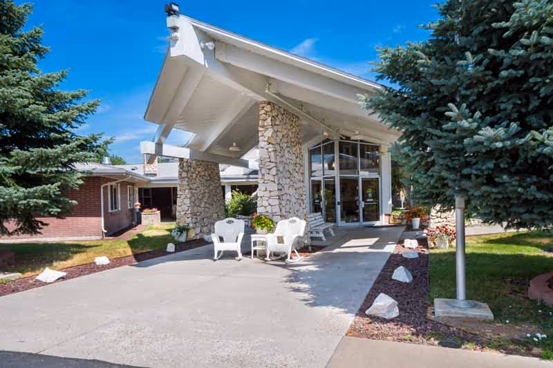 Front entrance of Elk Ridge Health and Rehabilitation Center with a covered stone-pillared porte-cochere, white chairs, and glass doors.