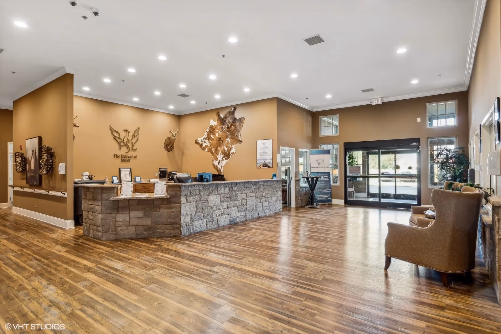Spacious senior living lobby with a stone reception desk, hardwood floors, seating area, and glass entry doors.