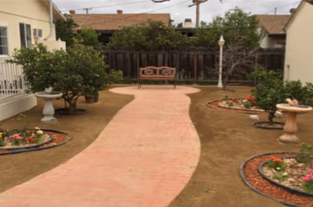 Curved pink brick pathway through a small courtyard garden with planted beds, birdbaths, a bench at the far end, and a lamp post.