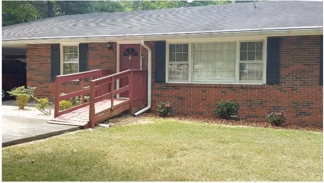 Exterior view of a single-story brick house with a gray shingled roof. There is a wooden ramp leading to a red front door with a small window. The house has two windows with white frames and black shutters. The lawn in front is green and well-maintained with small bushes near the house.