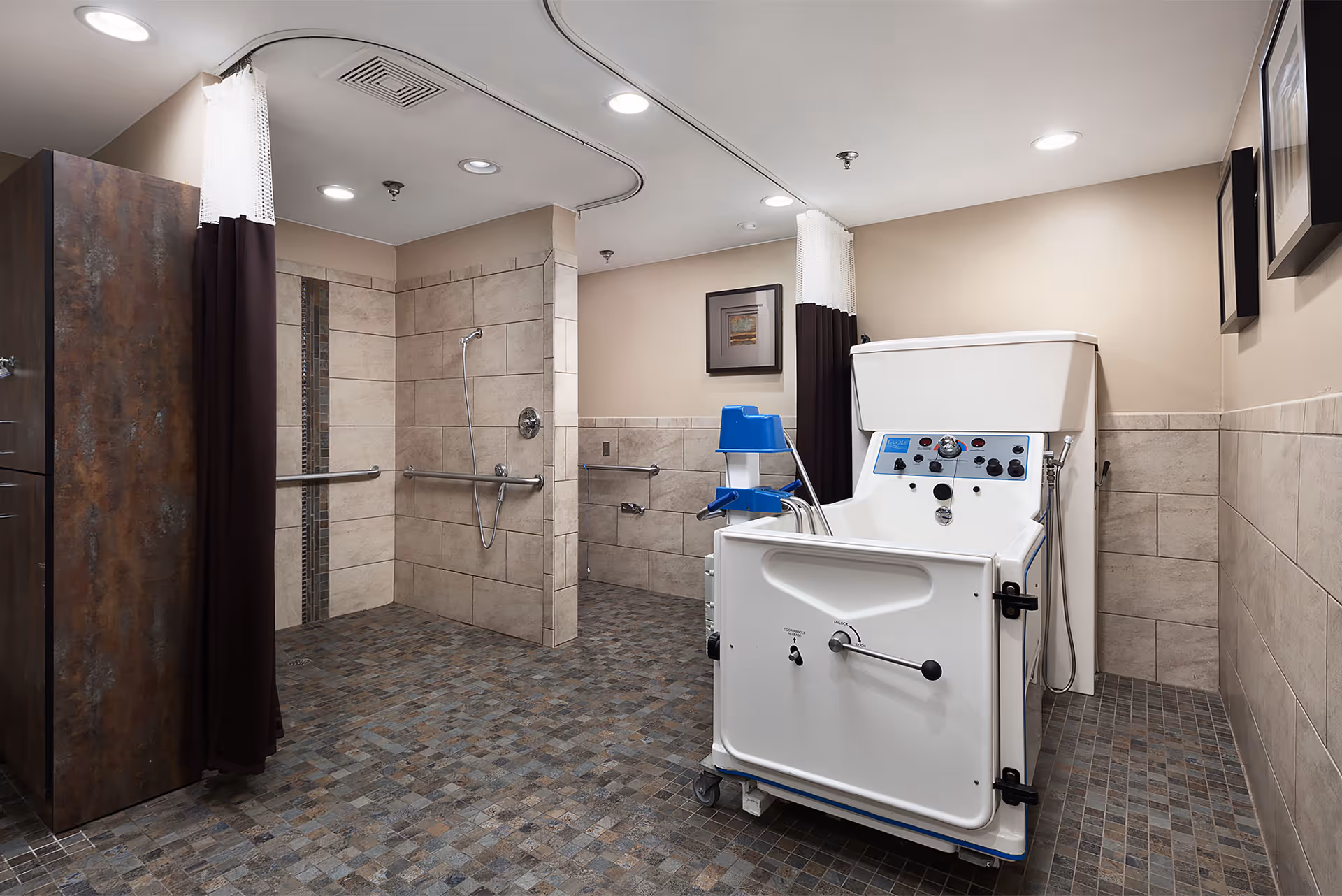 A spacious, accessible bathroom with tiled walls and floor, featuring a walk-in shower with grab bars and a handheld showerhead, a privacy curtain, and a large white bathing tub with control panel and handles designed for assisted bathing.